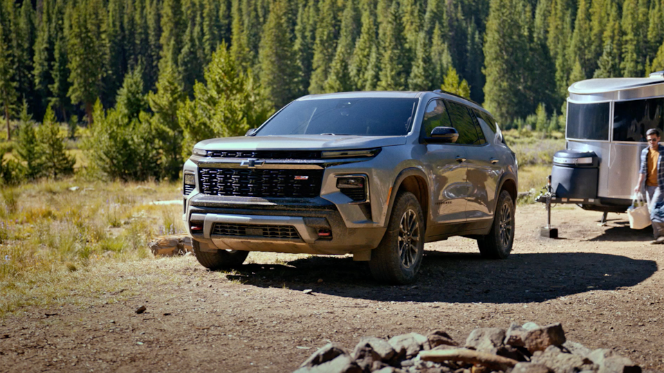 Front-side view of a 2025 Chevrolet Traverse SUV towing a trailer on a dirt path.