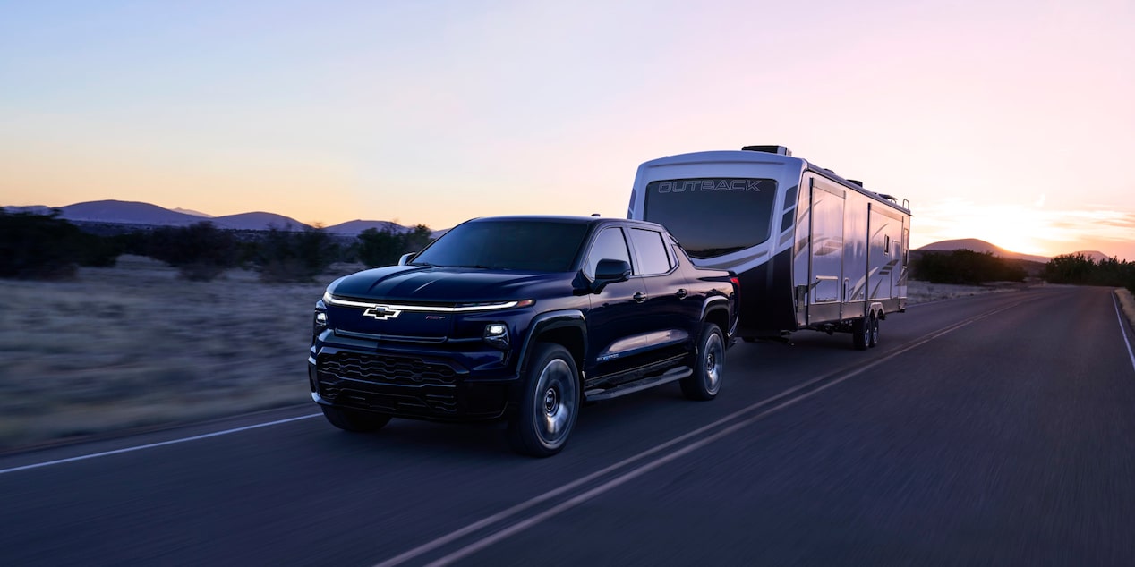 Front-side view of a 2025 Chevrolet Silverado EV with a trailer behind on a road at evening.