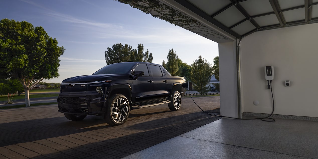 Un Chevrolet Silverado EV 2025 en train d’être rechargé à côté d’un garage pendant la journée.