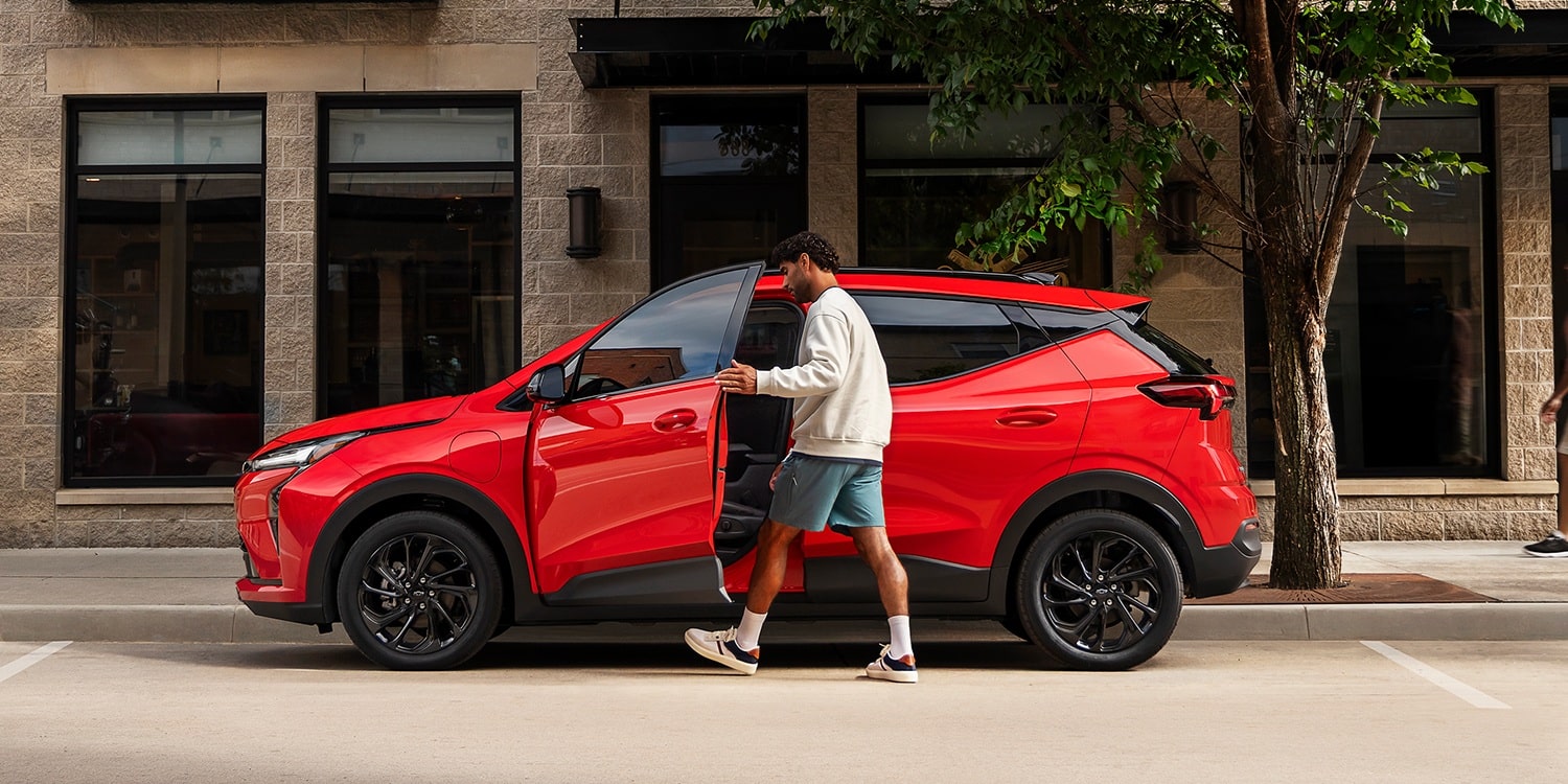 Vue latérale d’une Chevrolet Bolt 2027 rouge, avec un homme montant dans la portière du conducteur sur une rue de la ville.